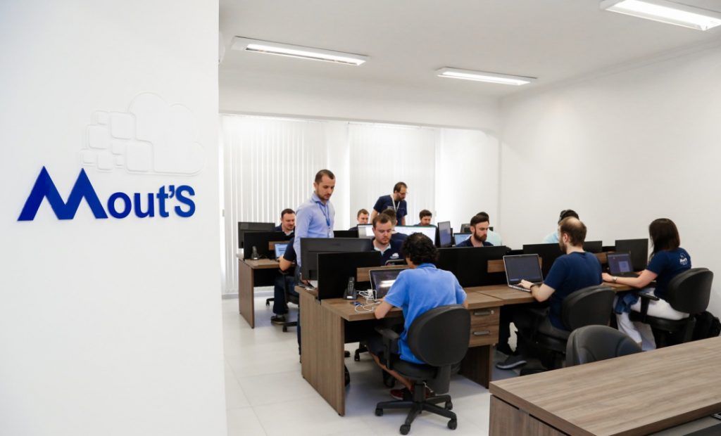 Imagem mostra sala da empresa Mout'S. Do lado esquerdo da imagem aparece a Logo da Empresa em uma parede branca, e do lado direito da imagem, aparece a sala com duas mesas grandes, com três pessoas de cada lado da mesa trabalhando nos computadores, e dois homens de pé ao lado das mesas.