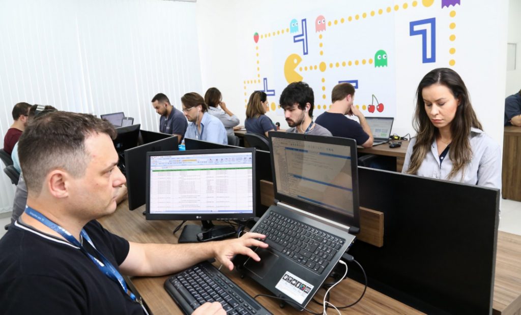 Imagem mostra sala de empresa com vários funcionários em suas cadeiras trabalhando em seus computadores.