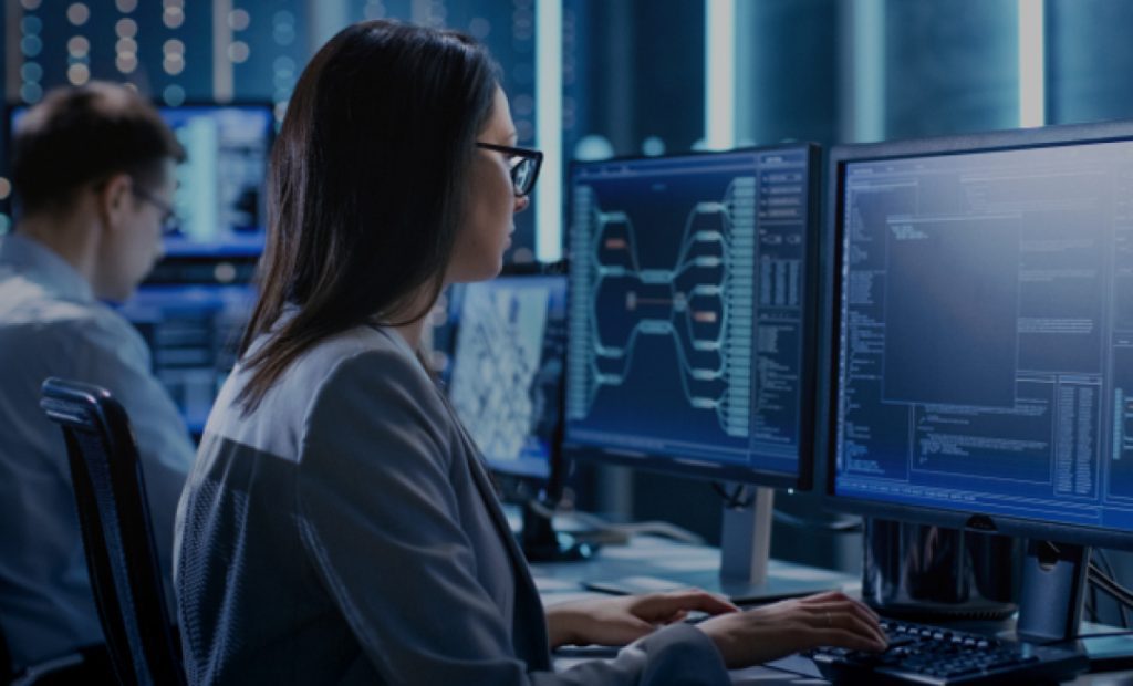 Imagem mostra uma mulher sentada de frente para uma mesa com um computador, mais atrás desfocado, esta outro homem também de frente para um computador.