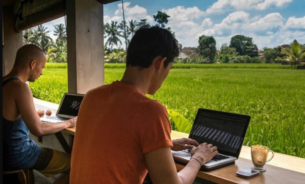 Foto de dois homens sentados trabalhando cada um com um notebook. A frente deles tem um vasto campo com grama e arvores no horizonte.