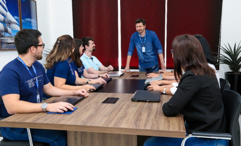 Imagem mostra uma sala de reunião, com um homem em pé em frente a mesa, quatro pessoas sentadas do lado esquerdo da mesa e outras três pessoas sentadas do lado direito.