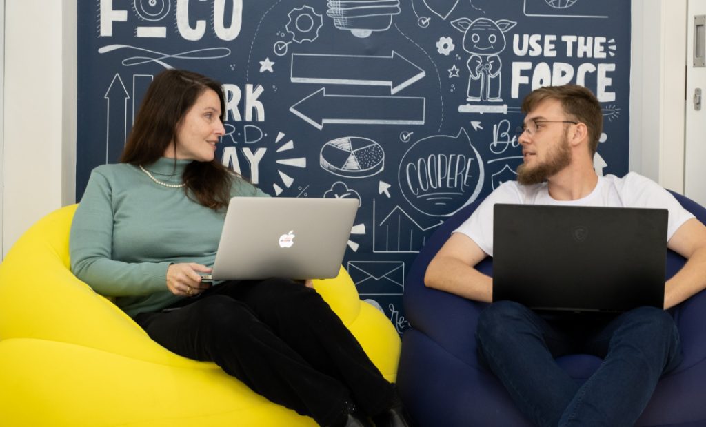 Foto de um homem e uma mulher sentados em puffs segurando cada um seu notebook e conversando.