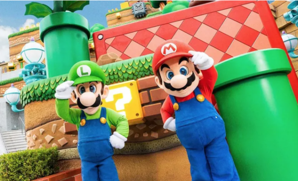 Imagem de dois personagens Super Mário fazendo pose para a câmera, de fundo tem o cenário do jogo bem colorido, o cenário tem forma circulares e retangulares com cores e texturas diferentes