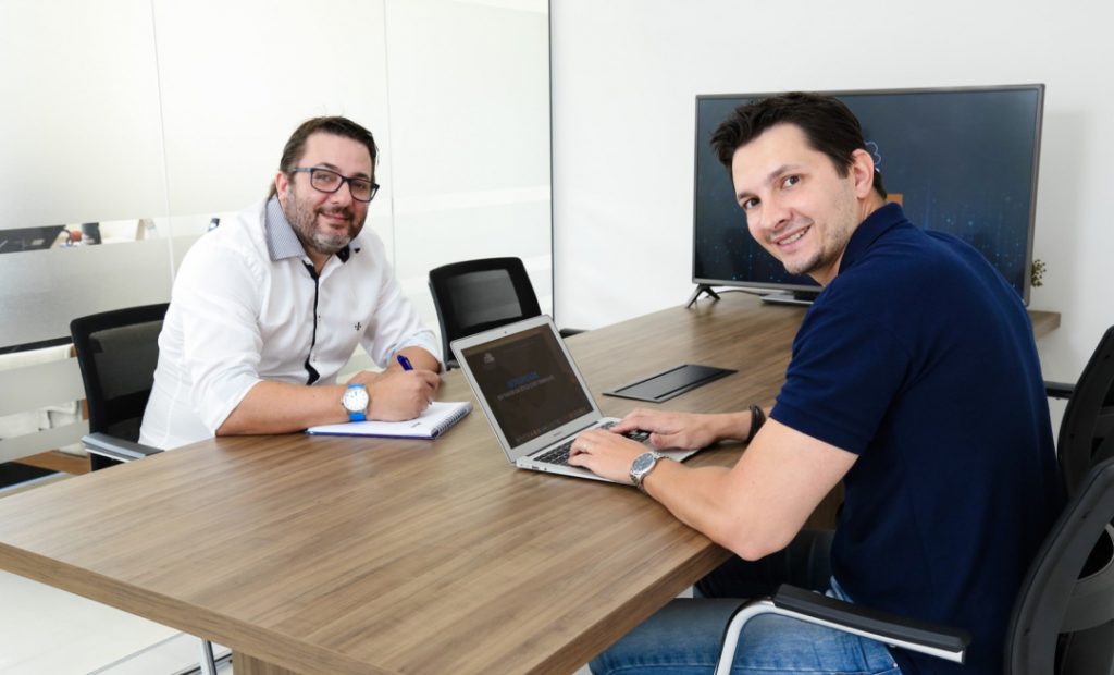 Imagem de dentro de uma sala de escritório, com dois homens sentados um a frente do outro sorrindo, um homem esta com um notebook a sua frente e outro esta com uma caneta na mão.