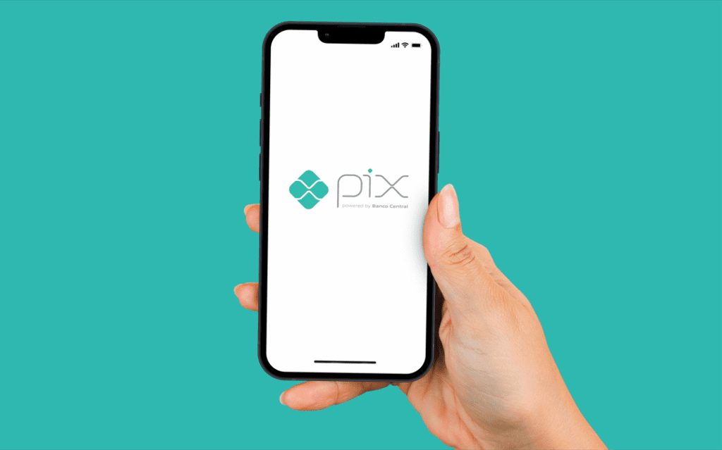 Imagem contém um fundo azul, na frente uma mão segurando um celular, na tela do celular aparece a logomarca do Pix.