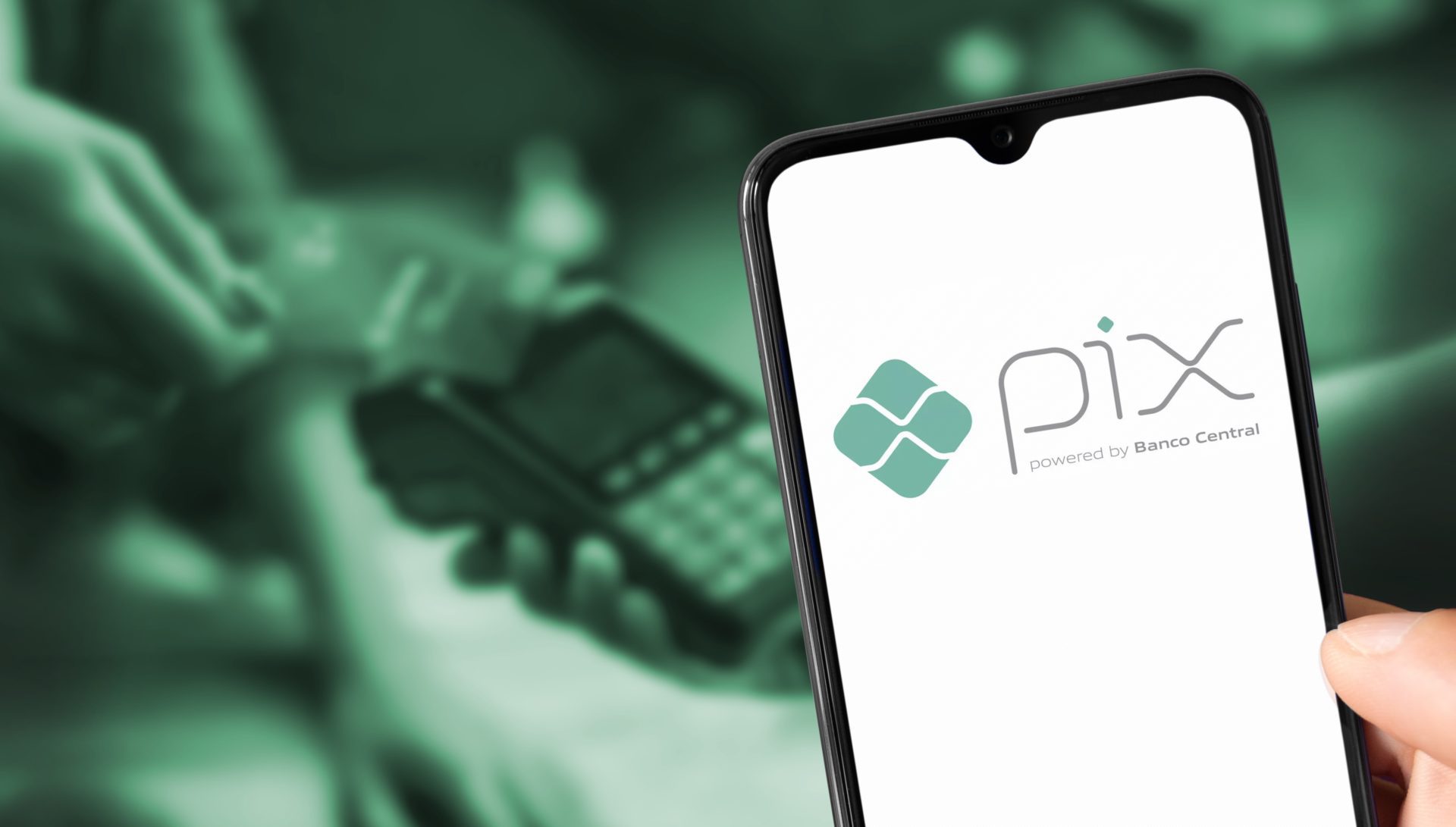 Imagem de fundo esta desfoca, mostrando uma pessoa passando cartão em uma maquininha de crédito, na frente da imagem aparece um celular com a logomarca do Pix.
